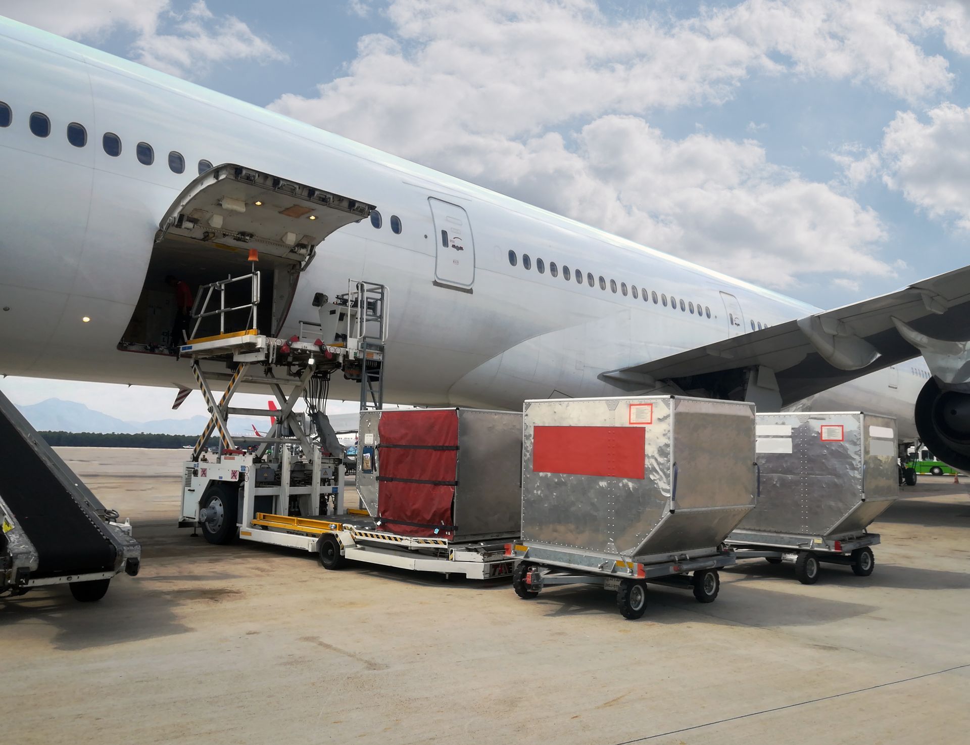 Contêineres de carga sendo carregados em um grande avião na pista, com um elevador de carga no compartimento de carga aberto.