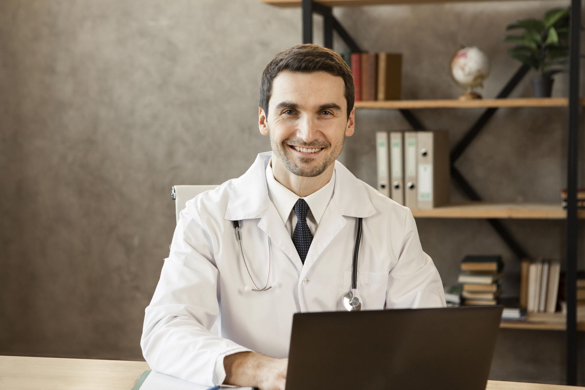 Doctor in a white coat smiling at a desk with a laptop in a clinic office