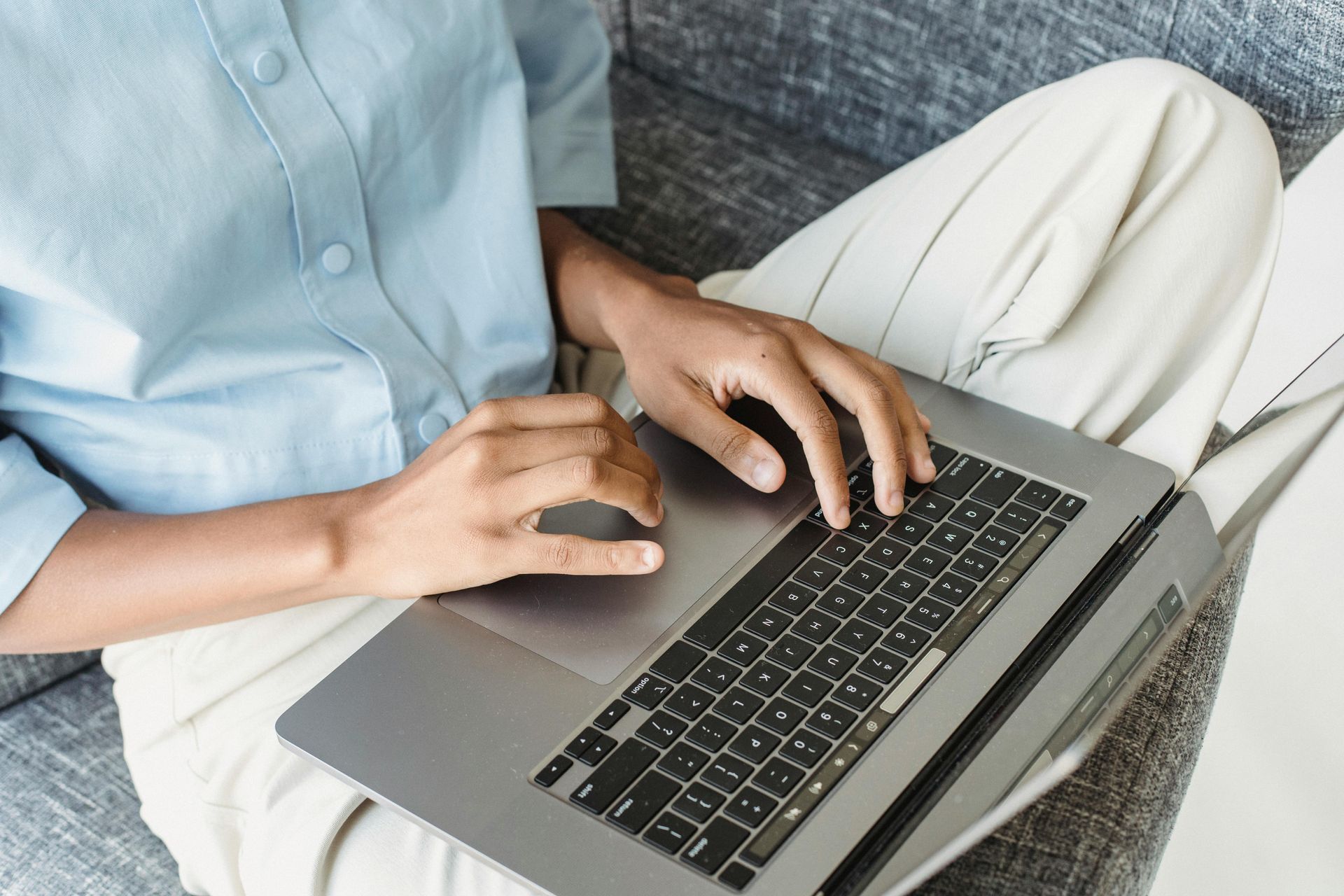 Pessoa digitando em um laptop sentada em um sofá branco, vestindo uma camisa azul-clara e calça creme.