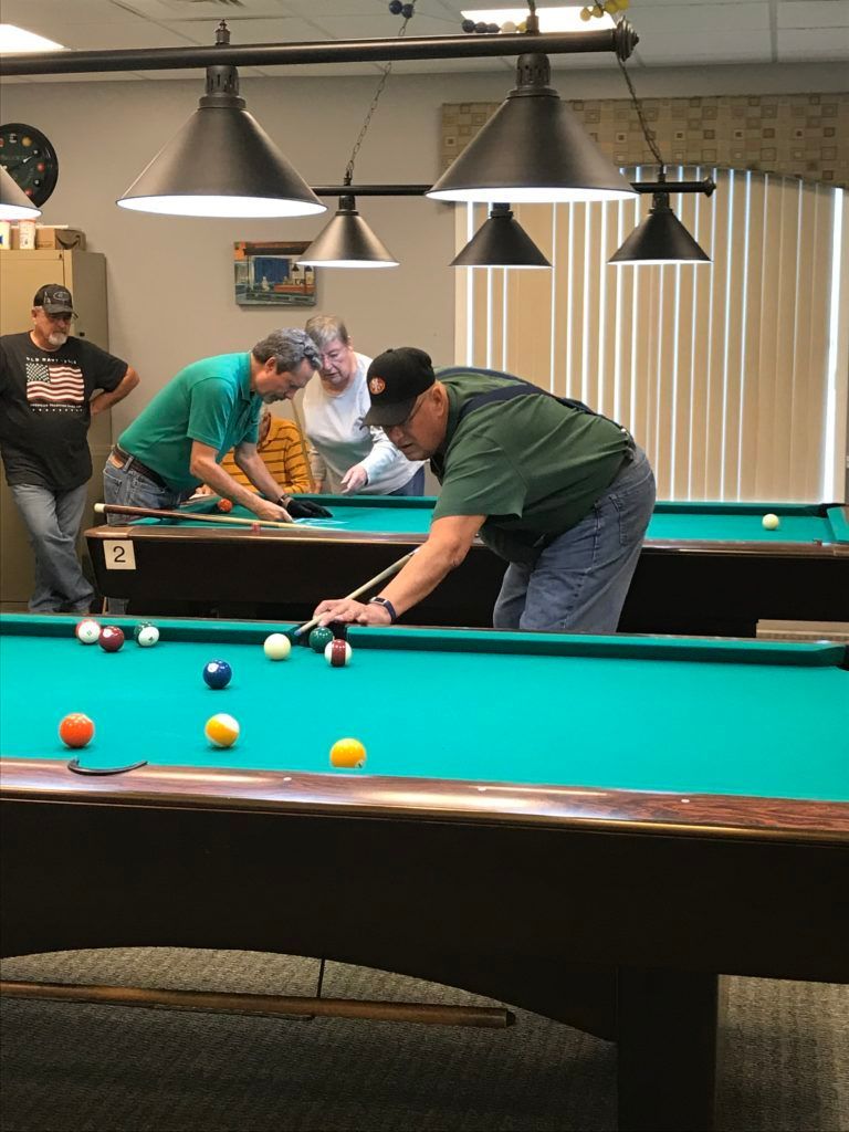 People playing pool around a green billiards table under hanging lamps in a game room