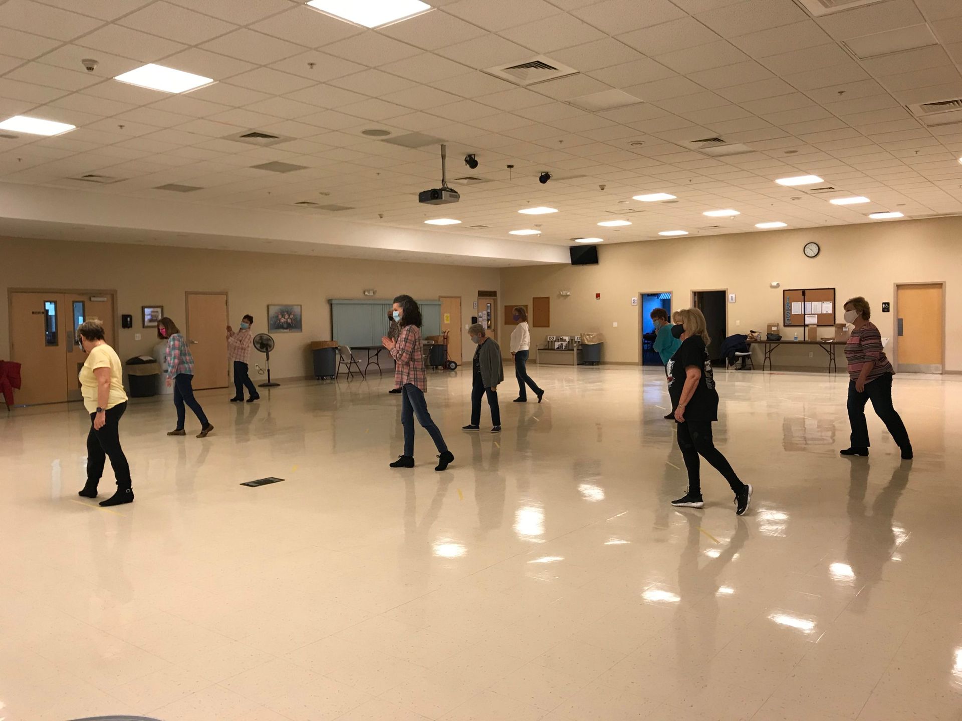 People dancing in a large beige community hall with polished floors and ceiling lights.