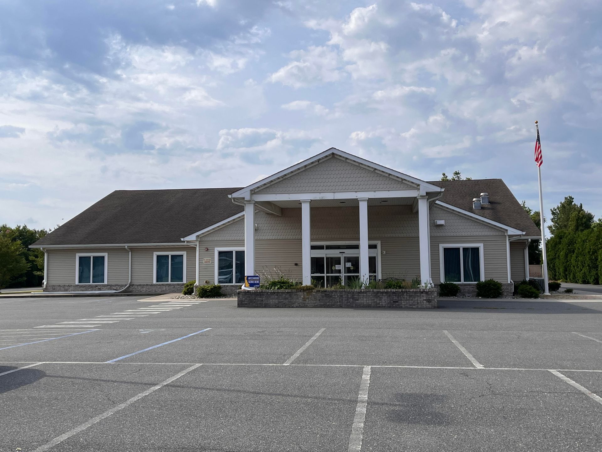 Low, light-gray civic building with white columns and an American flag in front of an empty parking lot