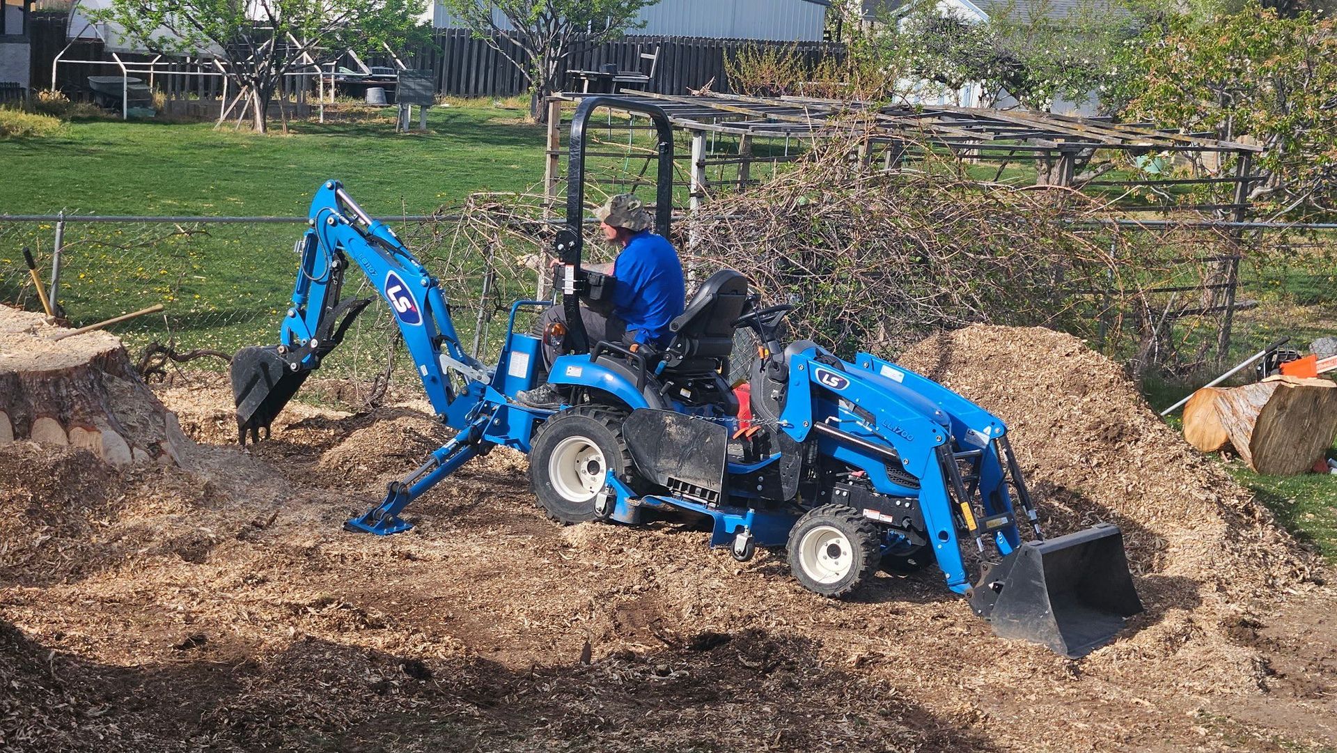 Blue backhoe loader working in a field, digging dirt near a fenced garden area.