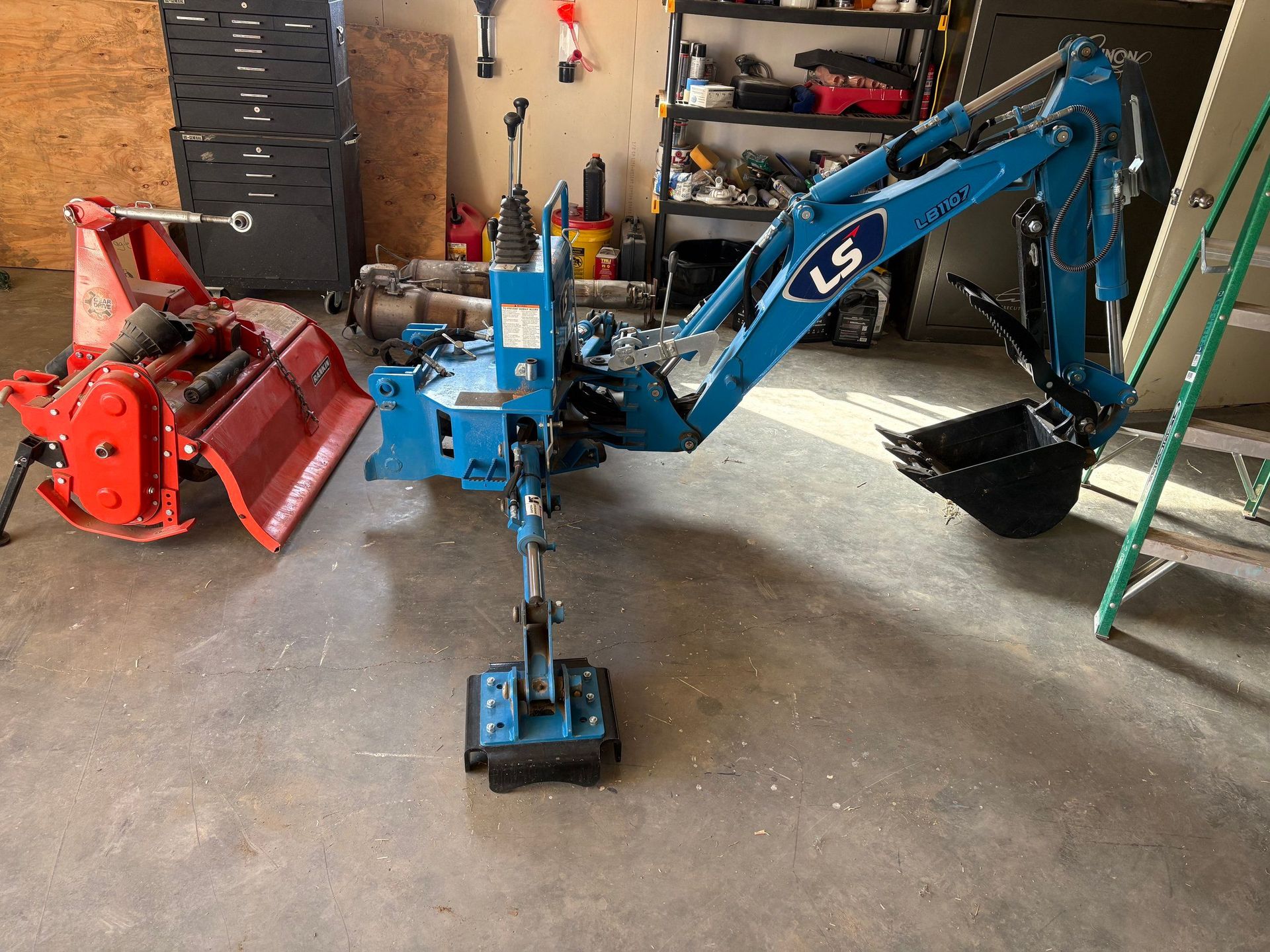 Small blue mini excavator with attachments in a garage, beside a red snow blower.