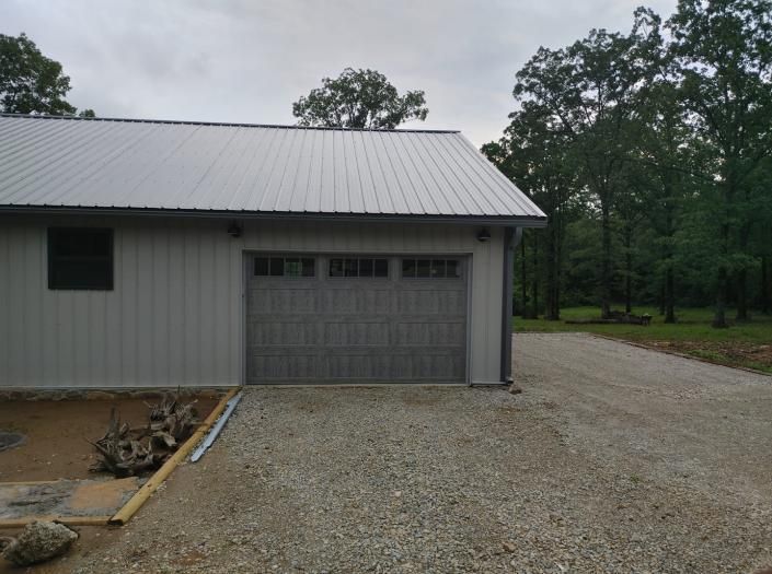 Gray metal building with a garage door beside a gravel driveway and trees