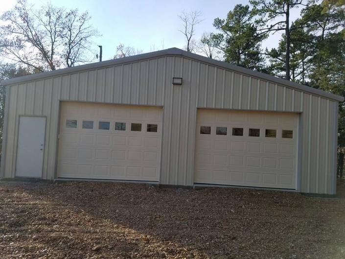 Two beige garage doors on a metal storage building with a side entry door, set on a wooded lot.