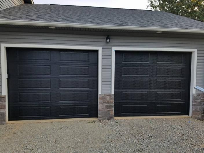 Two black garage doors on a house with gray siding and stone trim, above a gravel driveway