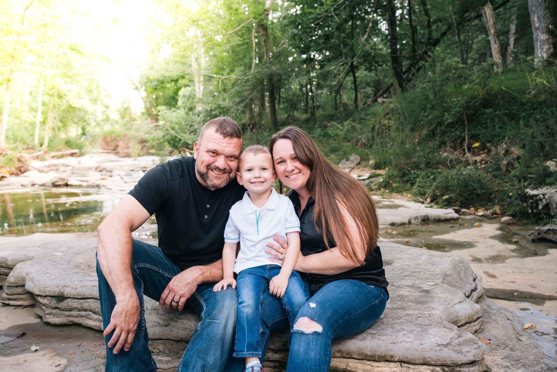 Family sitting on rocks by a creek in a wooded area, smiling at the camera.
