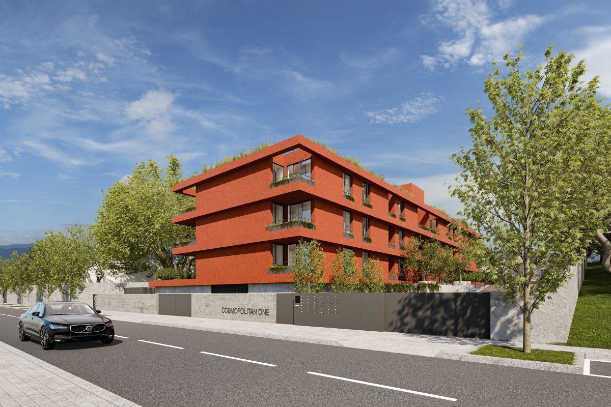 Prédio de apartamentos vermelho com varandas, um carro passando e árvores ladeando a rua sob um céu azul.