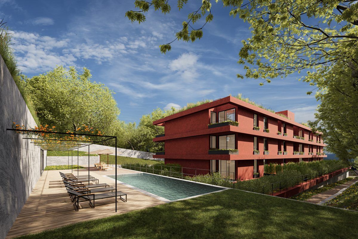 Edifício vermelho moderno com telhado verde perto de uma piscina e árvores, situado em uma encosta sob um céu azul.