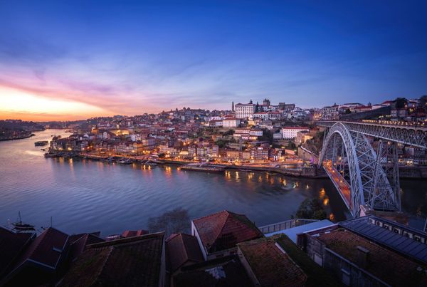 Paisagem urbana ao entardecer com um rio, uma ponte e prédios iluminados. Céu laranja e azul.