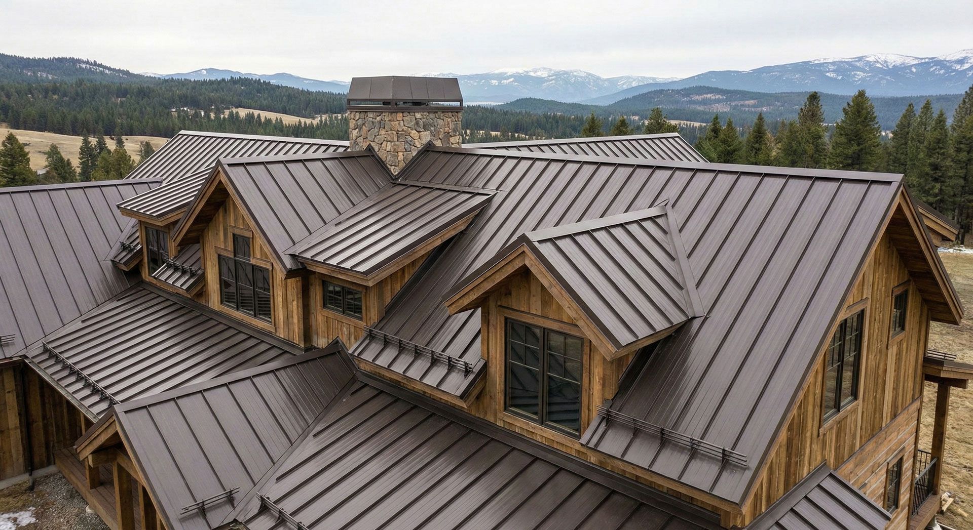 Mountain lodge with brown metal roofs and a stone chimney, surrounded by trees and distant hills