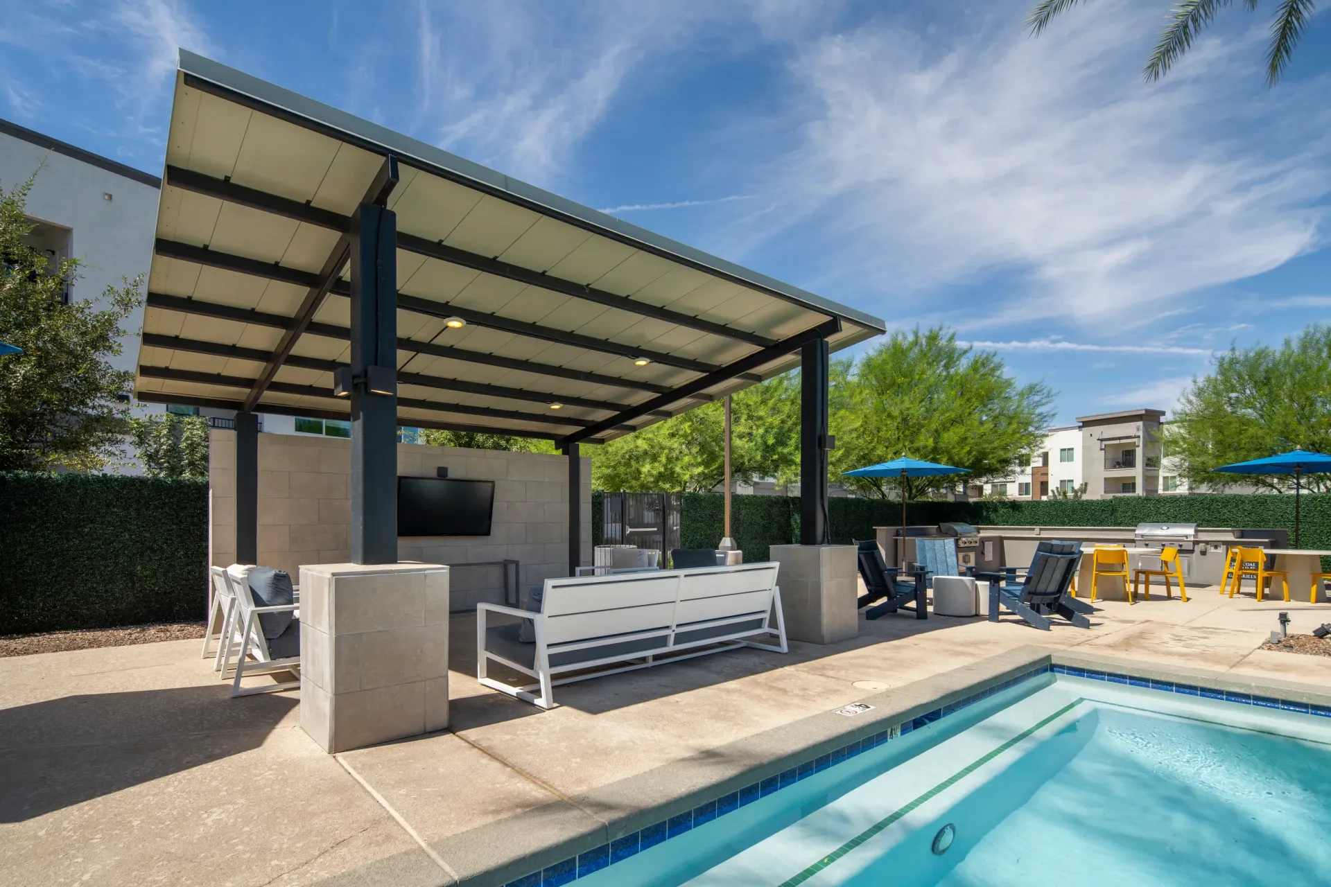 Outdoor covered lounge area with seating, TV, and grills next to a swimming pool at Lumina Mountain Vista in Mesa, AZ.