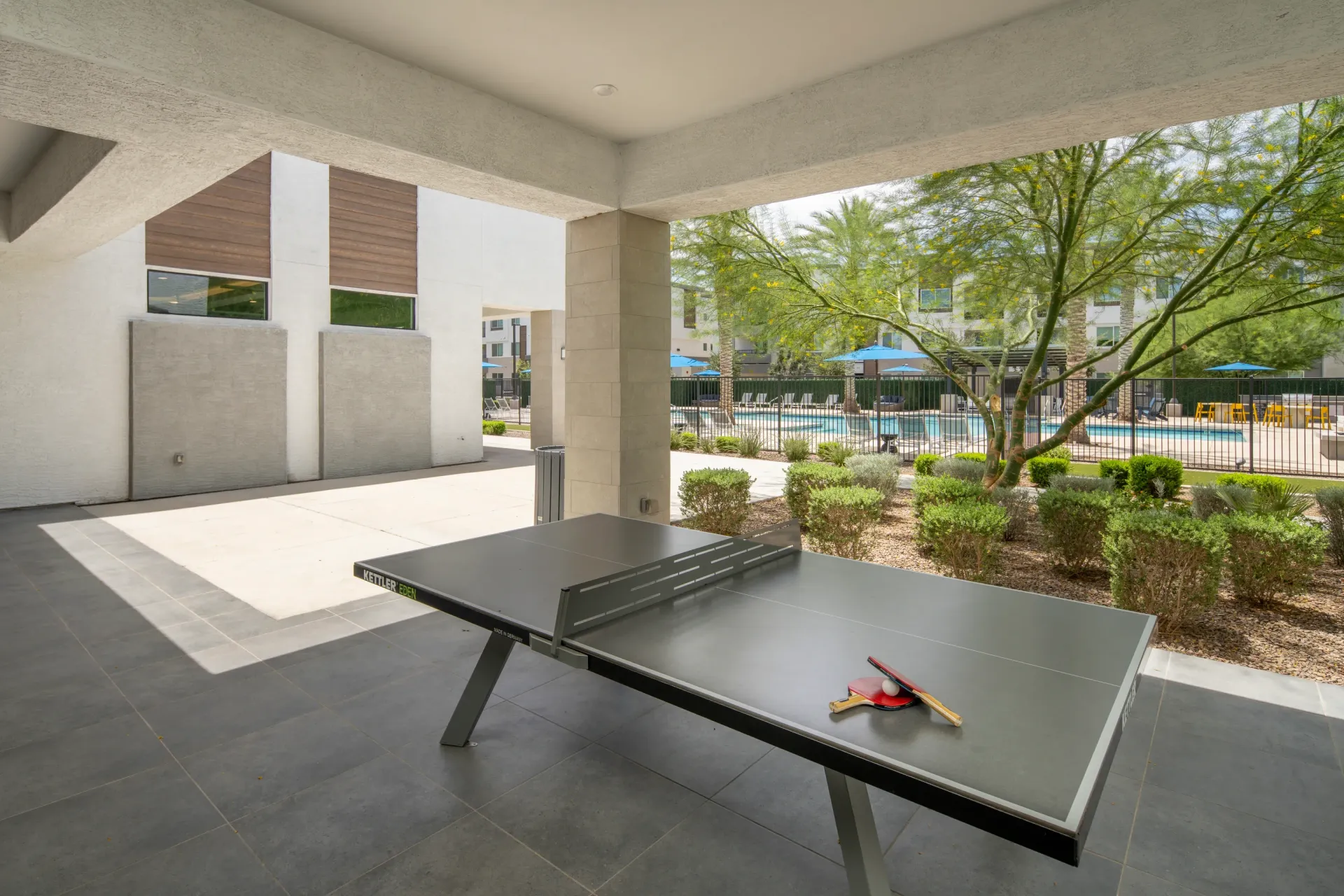 Ping pong table with paddles and balls in an outdoor covered patio area with a pool and lounge chairs in the background at Lumina Mountain Vista in Mesa, AZ.