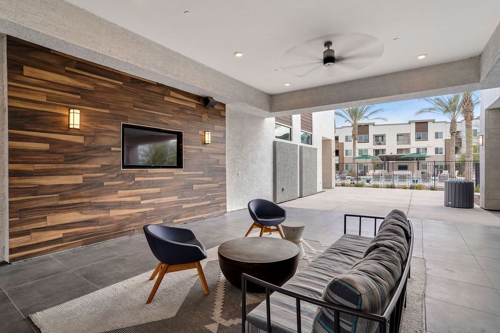 Covered outdoor lounge with wood-paneled accent wall, mounted TV, and seating; pool area visible in the background.