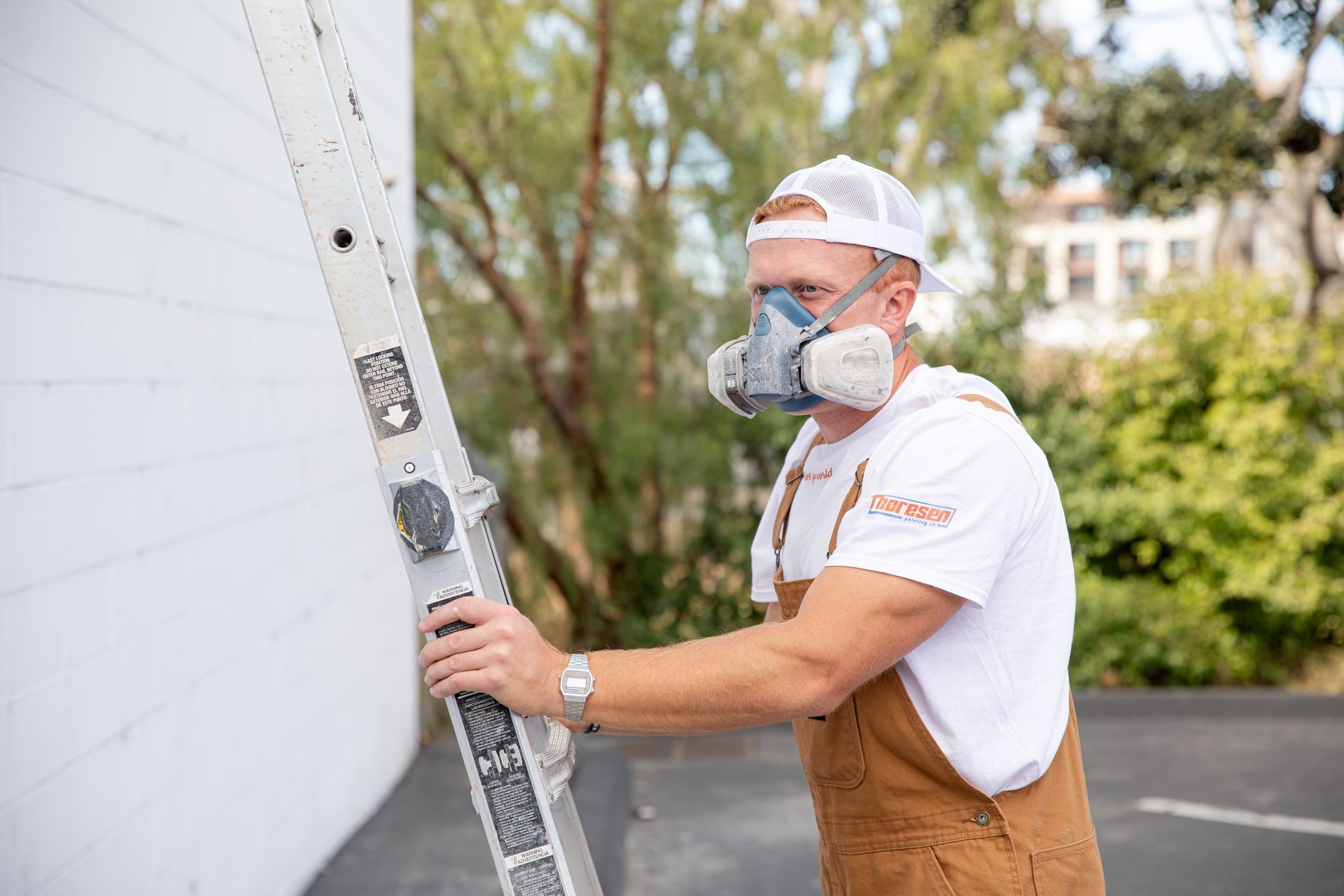 Man in respirator and overalls on ladder painting a white building exterior.