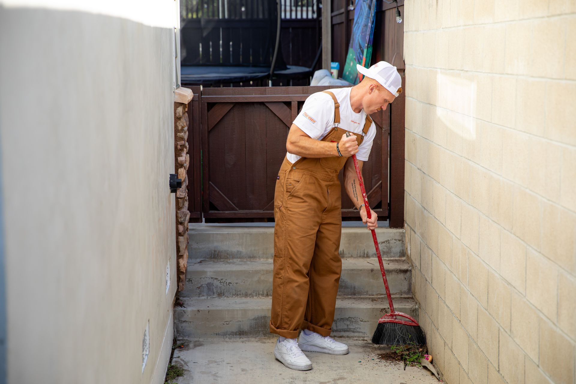 A man in overalls is sweeping the sidewalk with a broom.