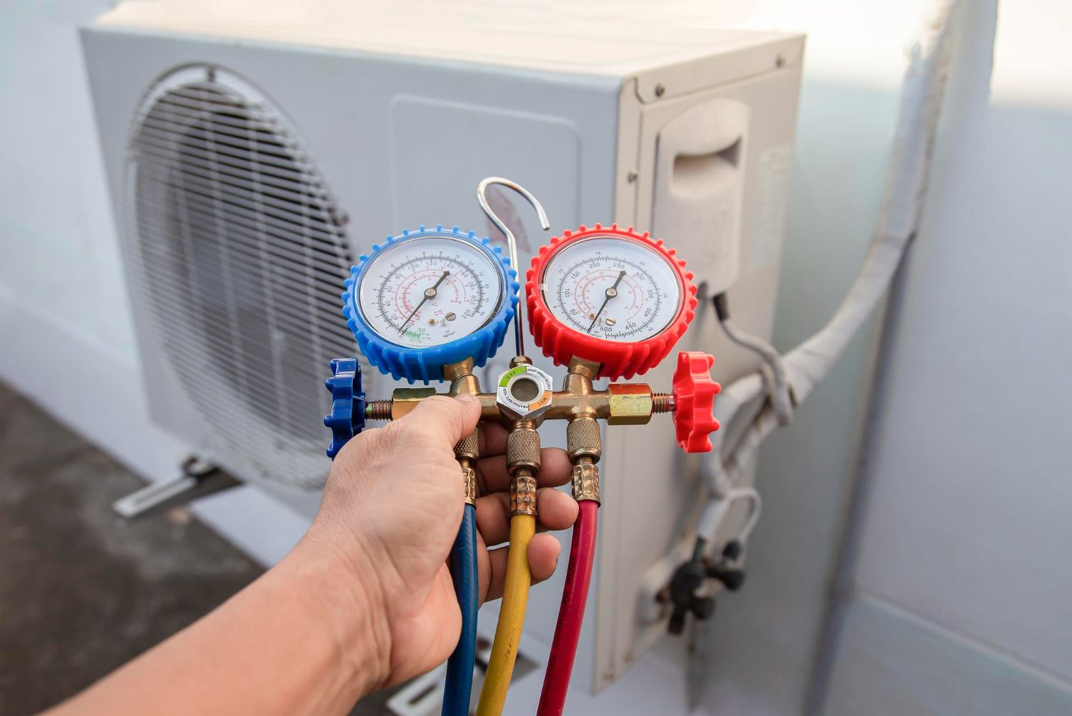 Hand holding HVAC manifold gauges connected to an outdoor air conditioner unit