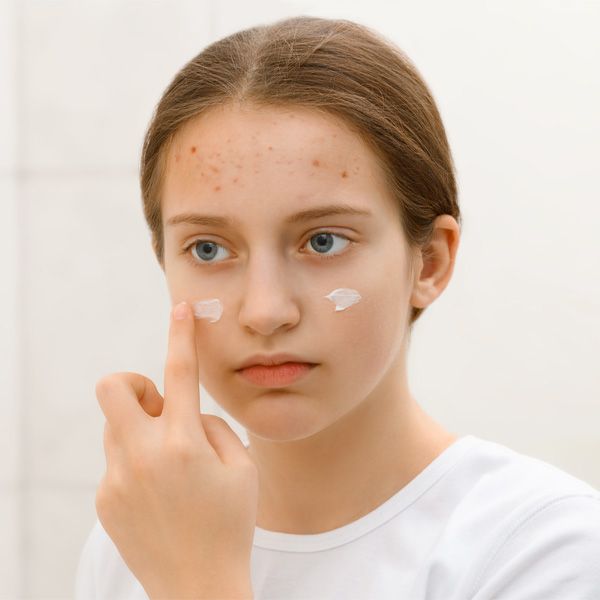 Teen applying white cream to cheeks in a bathroom mirror.