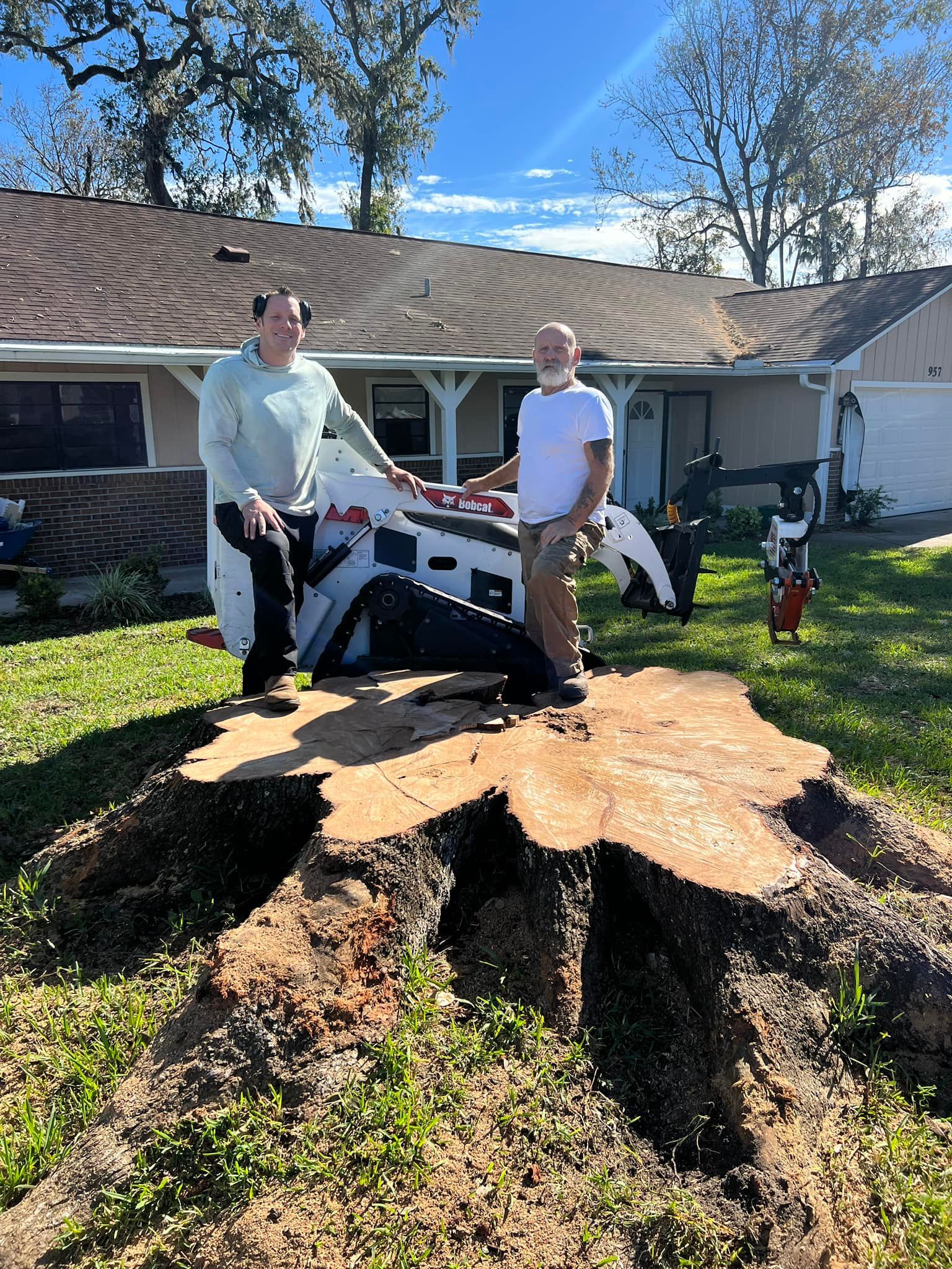 Two men are standing next to a large tree stump in front of a house.