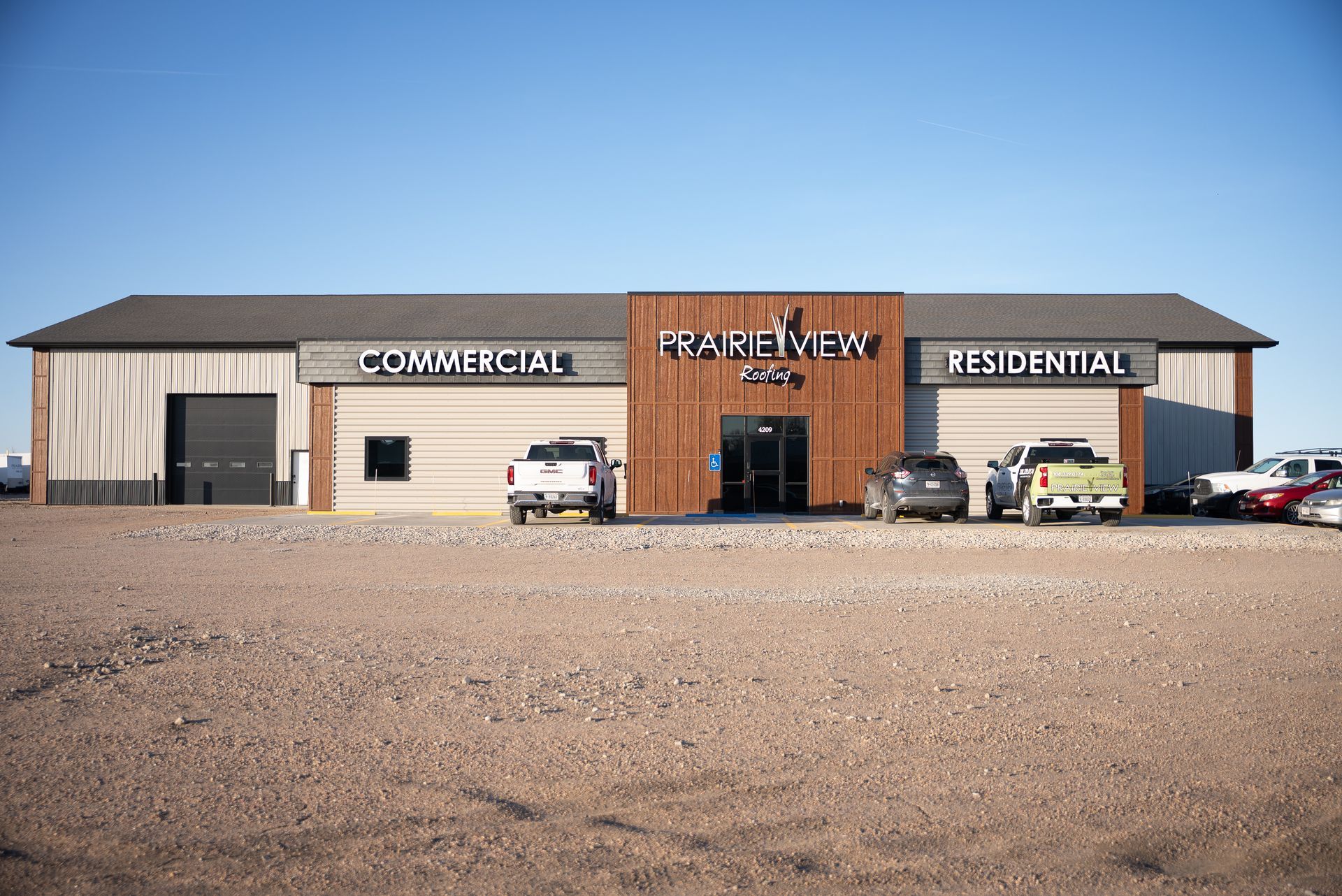 Small commercial building with separate commercial and residential sections, parked cars, and a gravel lot under a blue sky