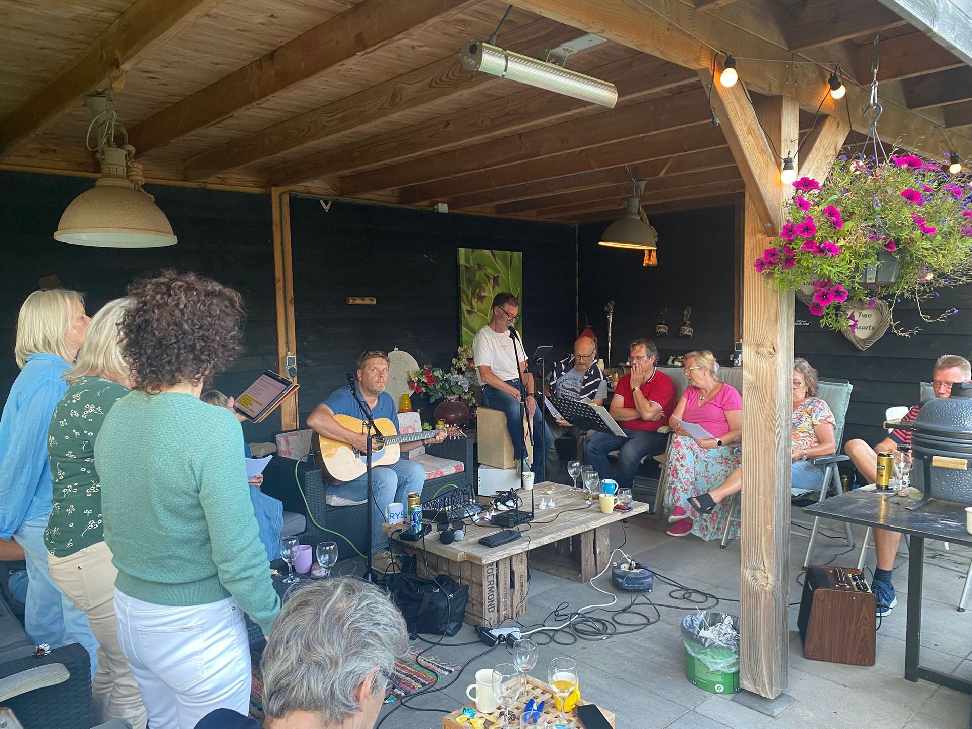 Mensen hadden zich verzameld onder een houten afdak en luisterden naar een man die gitaar speelde. Lichtslingers en bloemen waren zichtbaar.