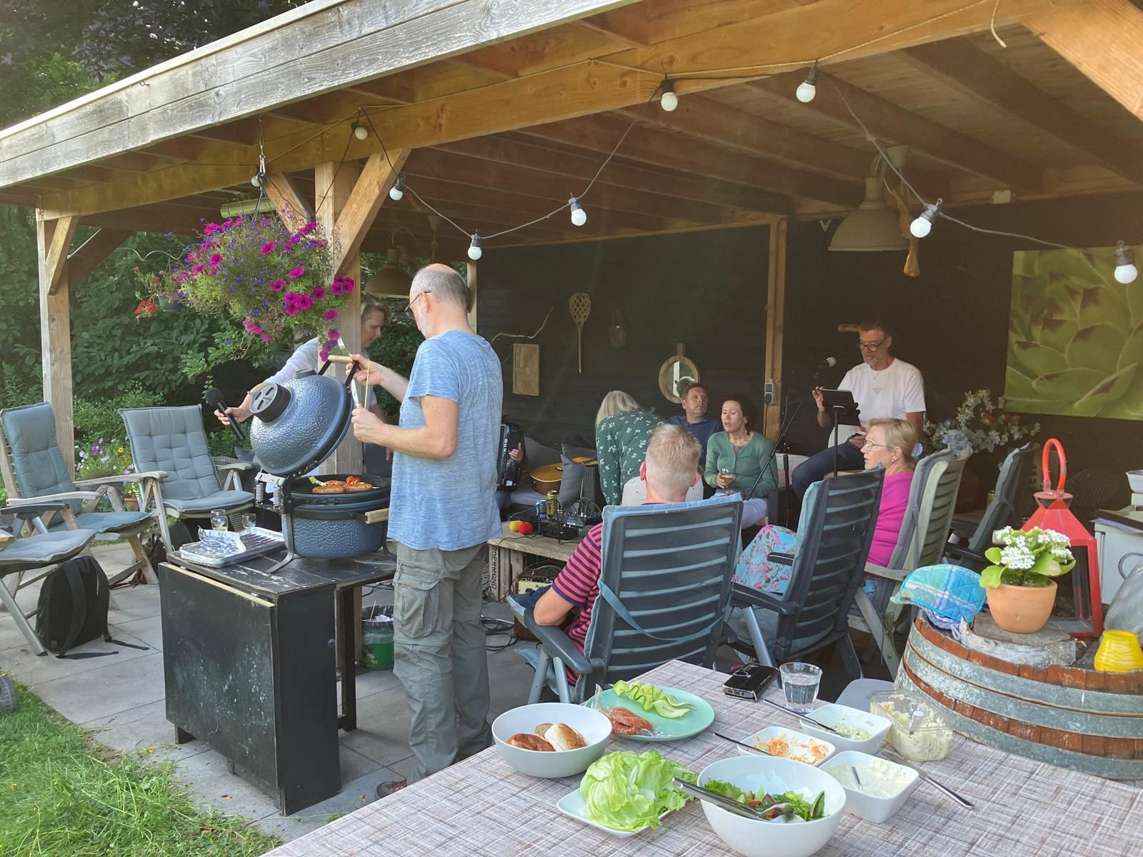 Mensen zitten bij een barbecue in de buitenlucht onder een houten afdak, er staat eten op tafel en er wordt gegrild.