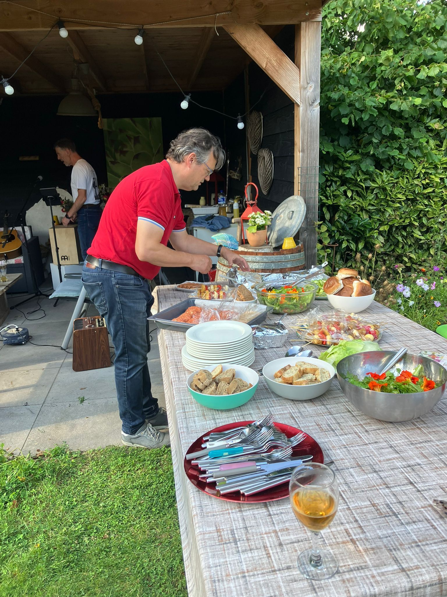 Een man in een rood shirt bereidt eten aan een buffettafel buiten; op de achtergrond is nog een persoon te zien.