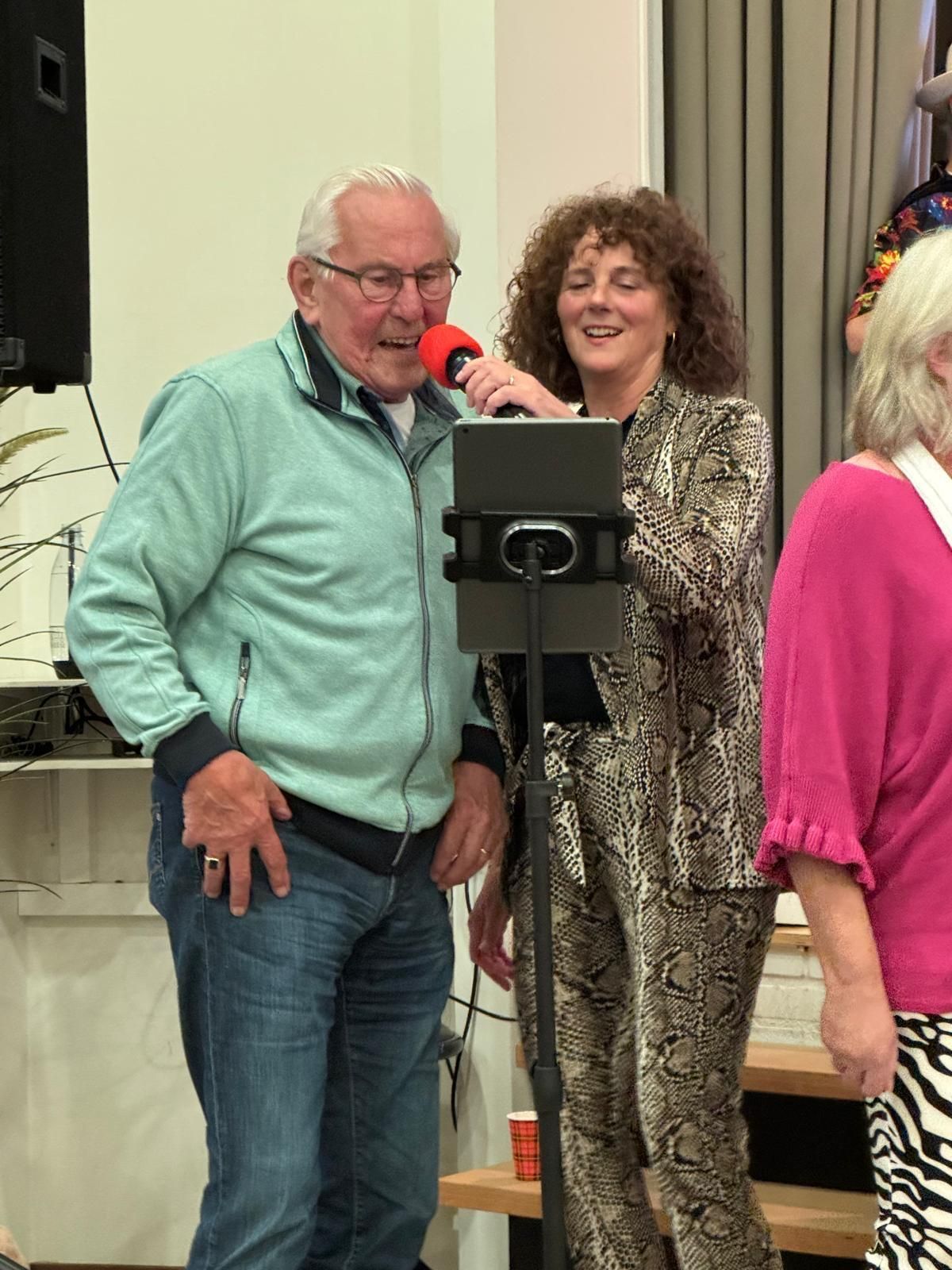 Een oudere man en een vrouw zingen samen in een microfoon tijdens een evenement; de vrouw glimlacht.