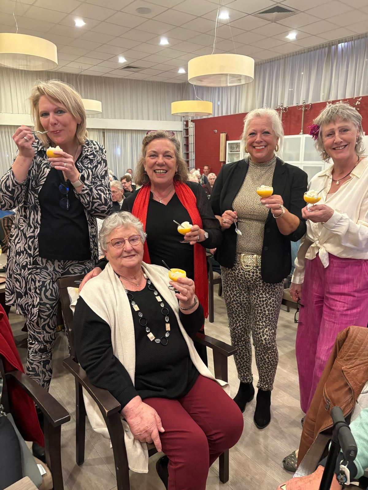 Vijf vrouwen zitten binnen en eten gele desserts. Sommigen dragen kleding met dierenprint, één heeft een rode sjaal om.