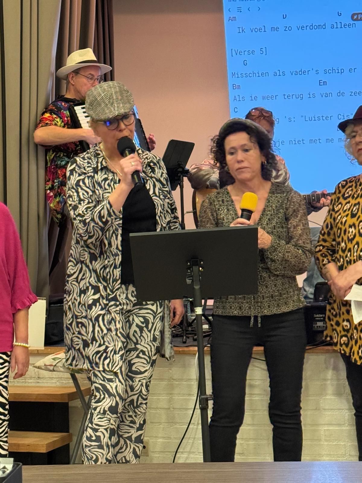 Groepszang, podium met band. Een vrouw in een pak met zebraprint zingt. Anderen houden microfoons vast. Podiumachtergrond, gedempte kleuren.