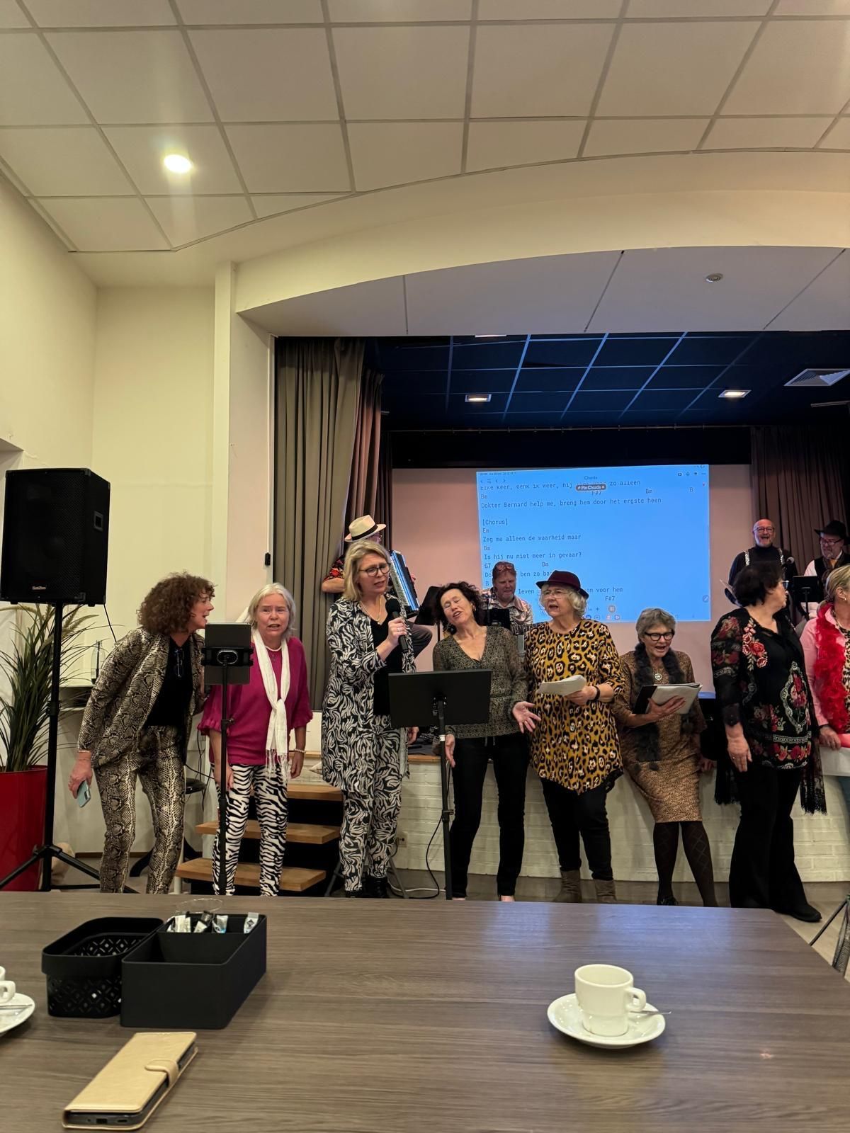 Een groep mensen op het podium, sommigen met microfoons, die een optreden geven in een zaal.