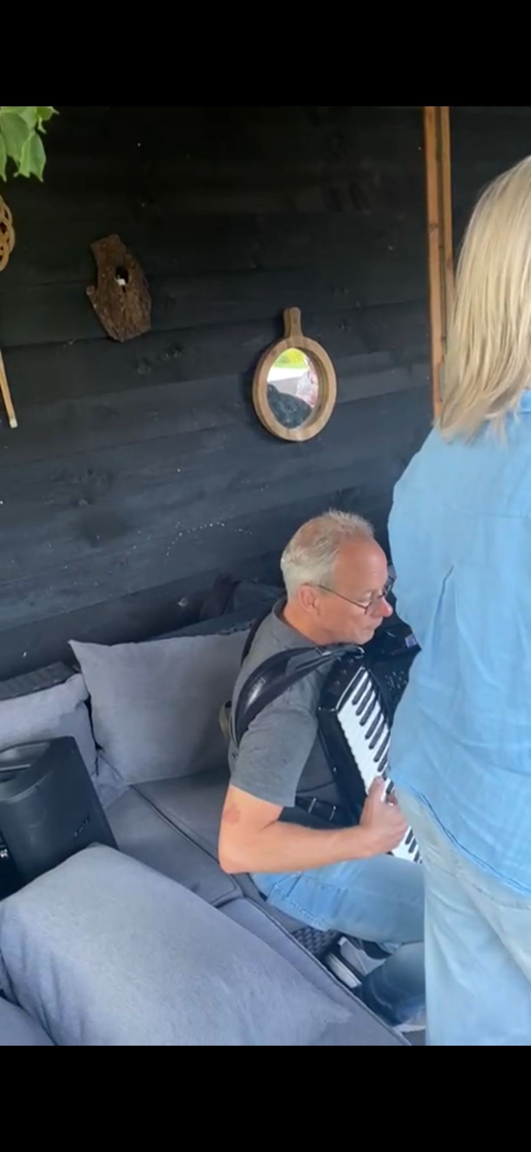 Een man speelt accordeon buiten, vlakbij een vrouw. Achter hen is een zwarte muur te zien, met grijze kussens op een bankje.