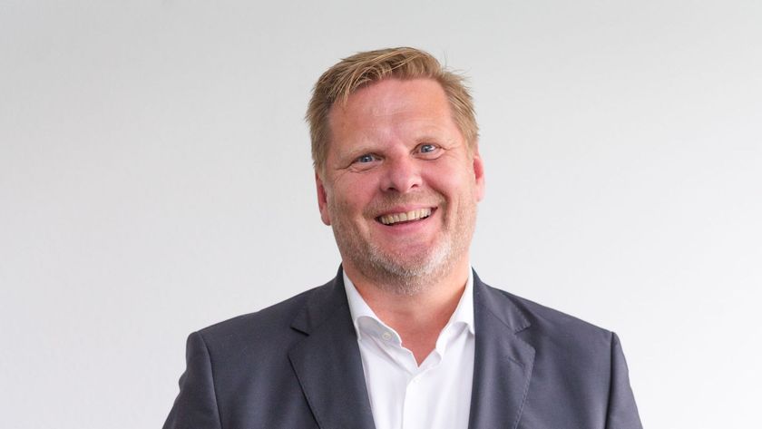 Een man met kort blond haar, gekleed in een wit overhemd en een donkerblauwe blazer, lacht breeduit tegen een witte achtergrond.