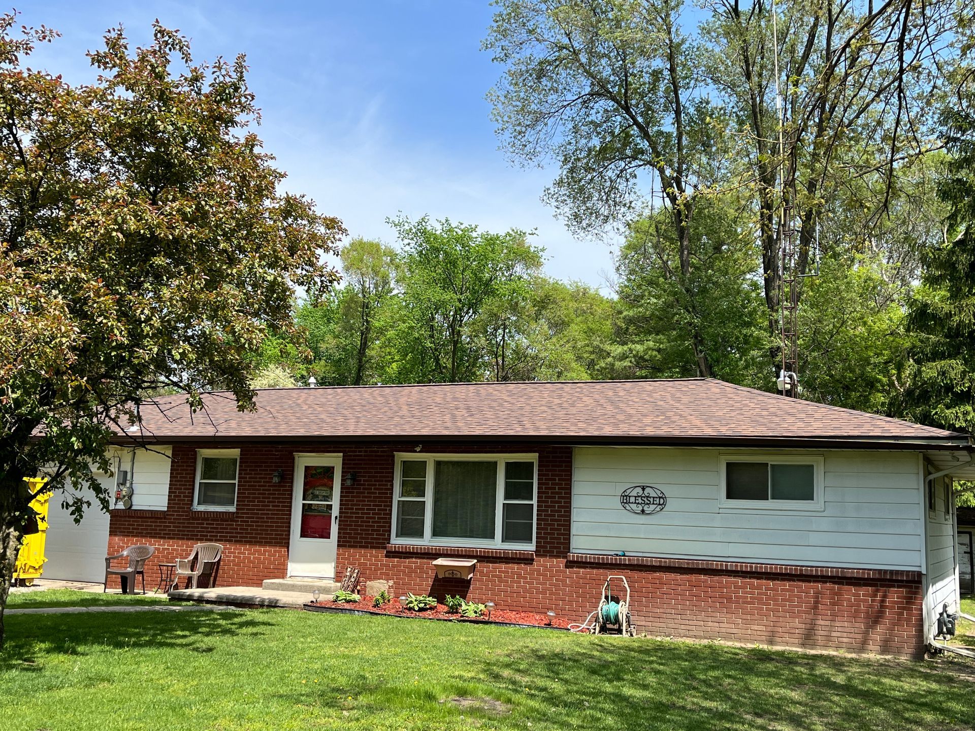 Ranch-style house with red brick and white siding, brown roof, surrounded by green grass and trees under a blue sky.