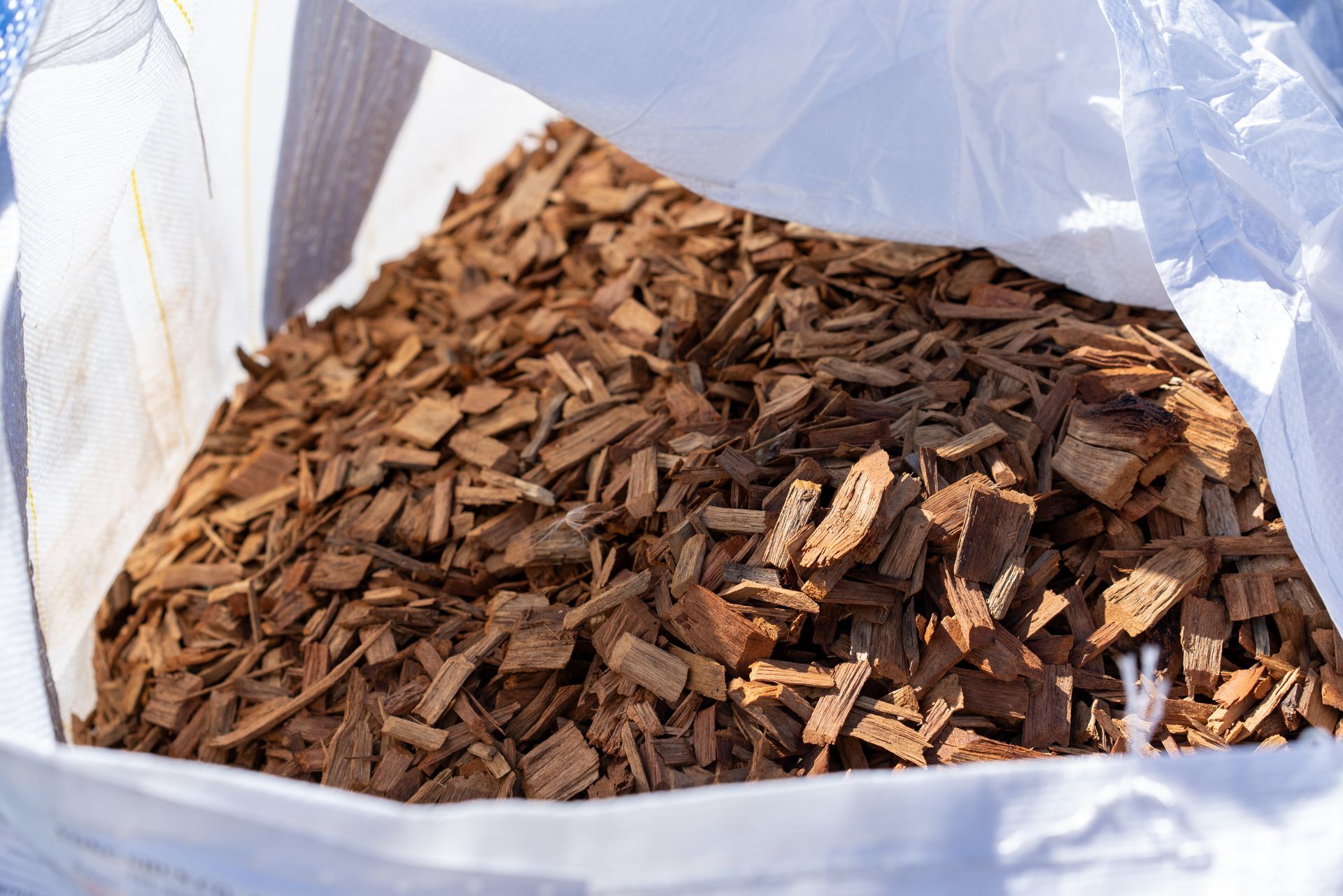 Brown wood chips mulch in large white bulk bag close-up view outdoors in sunlight.