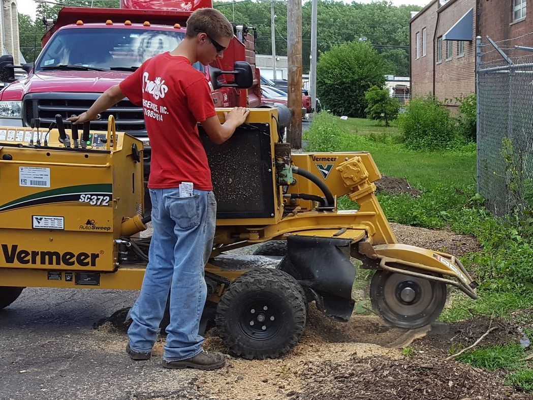 Worker operating a yellow Vermeer machine beside a red truck on a gravel roadside