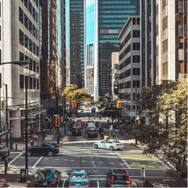 City street scene with tall buildings, cars, pedestrians, and traffic lights on a sunny day.