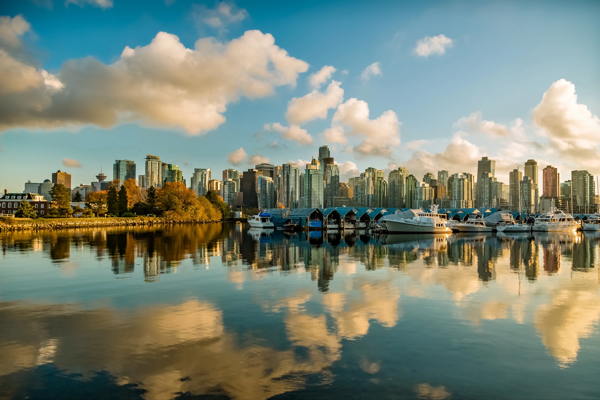 City skyline reflecting in calm water, boats docked, blue sky with clouds.