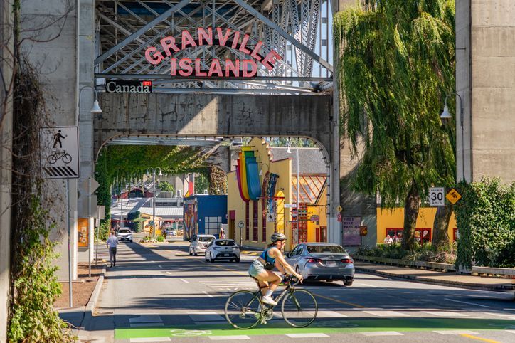 Cyclist on road entering Granville Island, Vancouver, Canada, under archway. Colorful buildings, signage, and trees.
