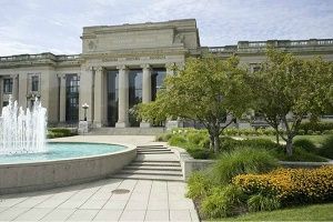 Exterior view of a museum with a fountain, trees, and landscaping in front. Building has columns and large windows.