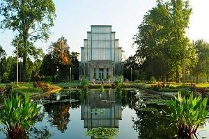 Glass building reflected in a pond, surrounded by lush greenery and trees.