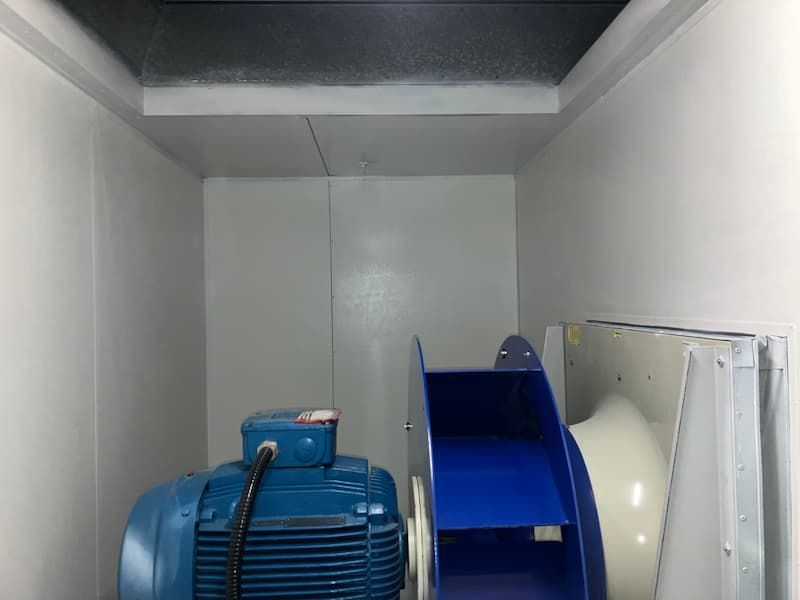 Inside a white-walled enclosure: blue motor, fan, and ductwork. Ceiling is dark.