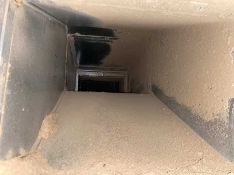 Inside view of a dusty, rectangular HVAC duct, showing layers of debris covering the metal surfaces.