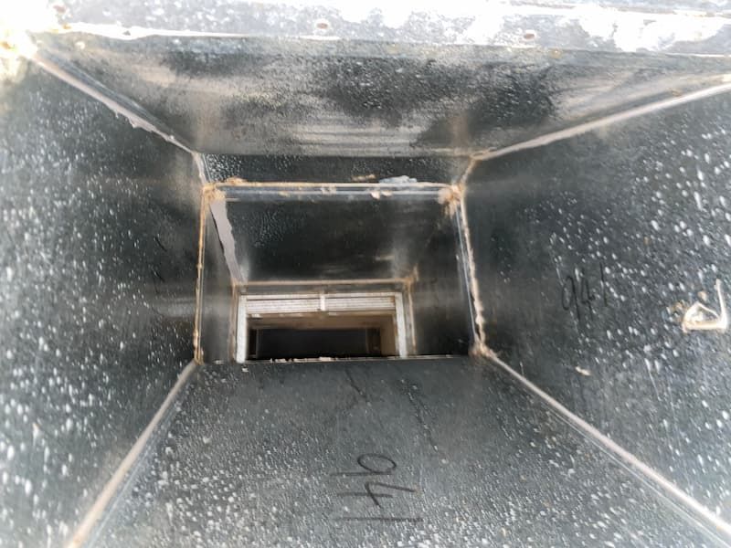 Inside a rectangular metal duct, viewed from an opening, with gray dusty walls and a rectangular opening at the end.
