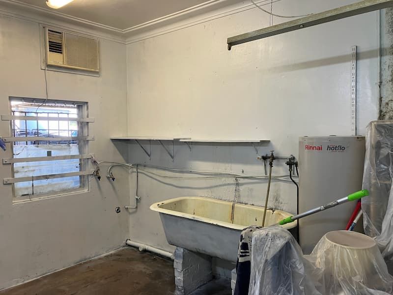 White-walled room with a window, shelves, tub, and water heater. A/C unit and utility items present.
