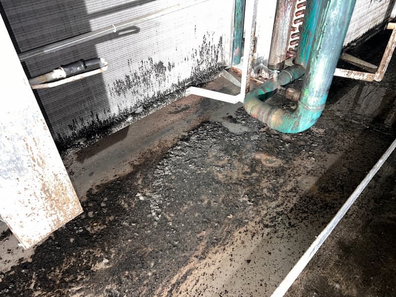 Black debris and water accumulate around AC unit piping on a rooftop.