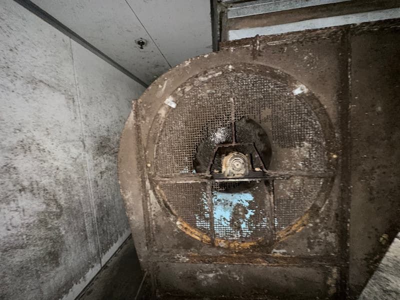 Dirty industrial fan covered in dust and debris, located in a narrow, gray enclosure.