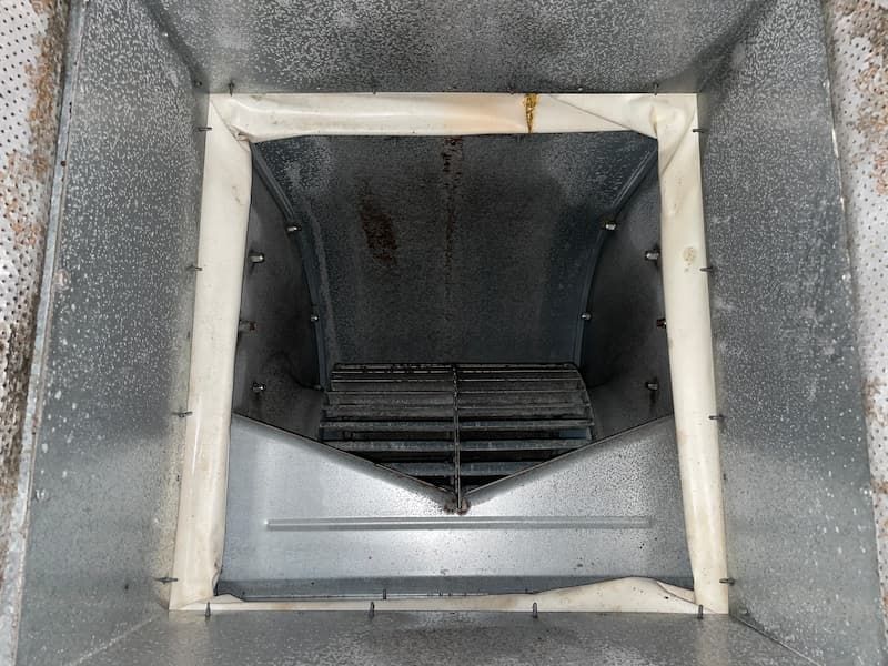 Inside of a metal air duct with a filter and fan, dirty with some debris.