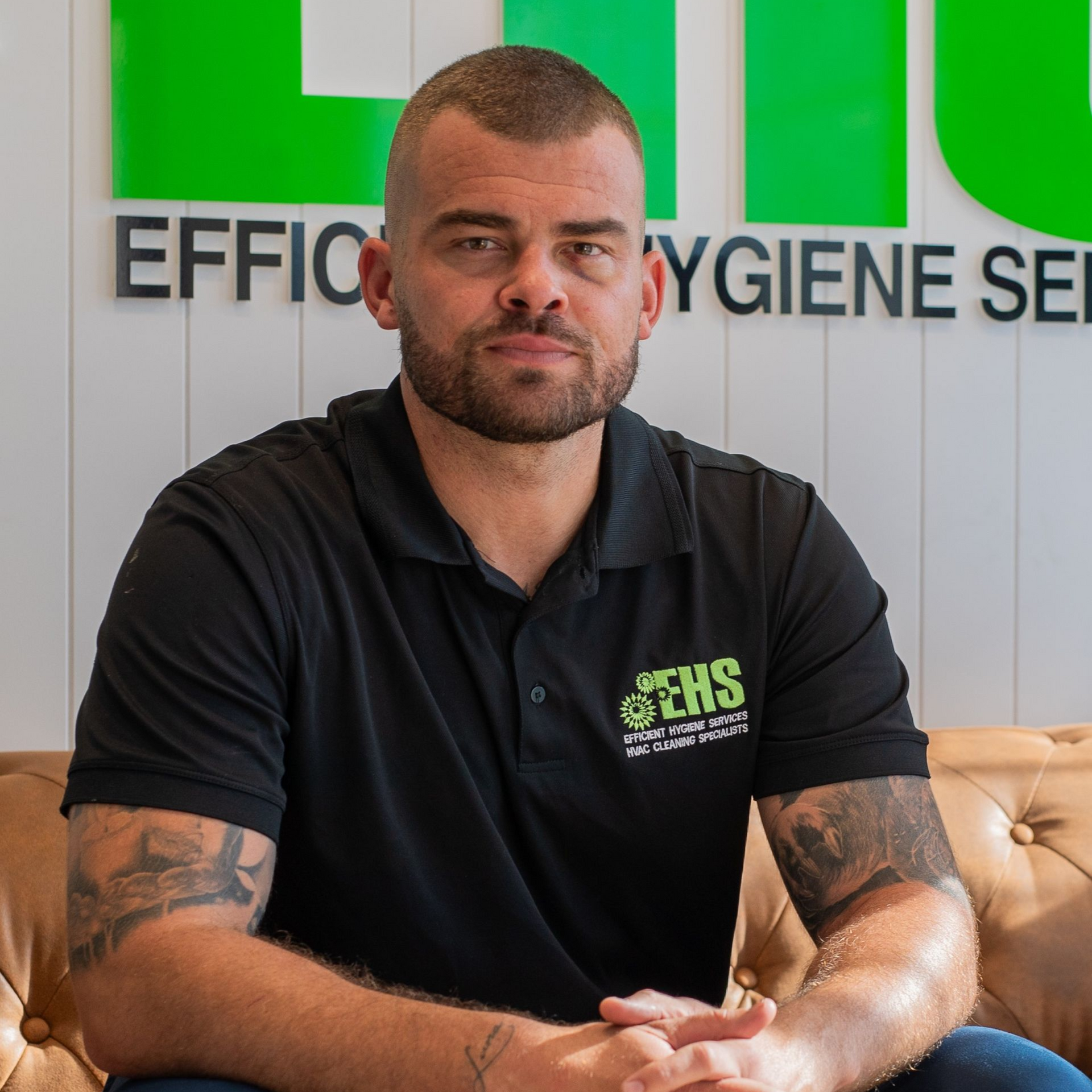 Man with a shaved head and tattoos sits on a brown couch; green and white logo in background.